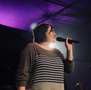 Woman in a gray and white striped color block shirt singing into a microphone on a stage with purple backlights.